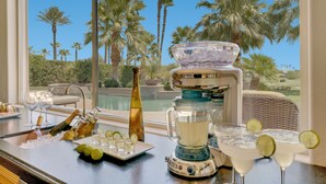 A bar area with a view of palm trees and a pool, featuring a blender, glasses, and bottles. - FALL GETAWAYS 25% OFF - Private and Serene - Room for the whole Family (La Quinta)