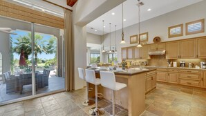 A modern kitchen with a large island, bar stools, and a dining area with a glass door overlooking a garden. - FALL GETAWAYS 25% OFF - Private and Serene - Room for the whole Family (La Quinta)