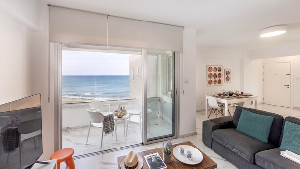 Ein modernes Wohnzimmer mit einem großen Schiebeglasfenster, das auf einen Balkon mit Meerblick führt. Es gibt ein Sofa, einen Couchtisch und links einen Fernseher.