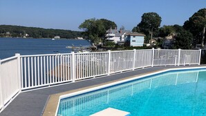 A swimming pool with a diving board, surrounded by a white fence, with a view of a lake and houses in the background. - Direct waterfront vacation rental, family friendly, pets allowed (East Lyme)