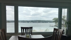 A dining area with a table and chairs facing a large window overlooking a body of water with boats and a distant shoreline. - Direct waterfront vacation rental, family friendly, pets allowed (East Lyme)