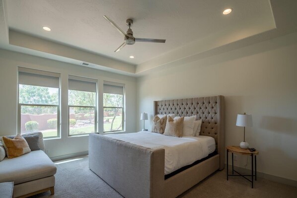 A bedroom with a large bed, a ceiling fan, and a view of the outdoors through large windows.
