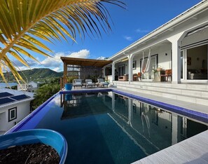 Ein Schwimmbecken mit Liegestühlen, einer überdachten Terrasse und Blick auf die Berge.