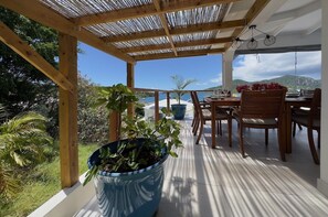 Ein überdachter Außenbereich zum Essen mit Blick auf das Meer, Holzmöbel und Kübelpflanzen.