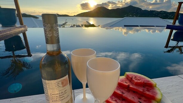 Pöydällä on pullo väkevää alkoholijuomaa, kaksi viinilasia ja lautanen vesimeloninviipaleita. Taustalla näkyy vesistö ja vuoristo.