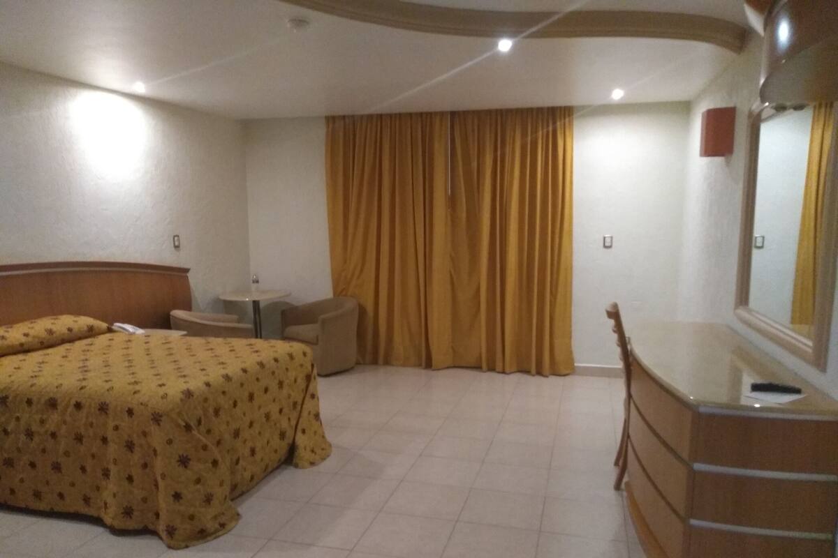 A hotel room with a bed, a desk, a chair, a dresser, and a large window with curtains.