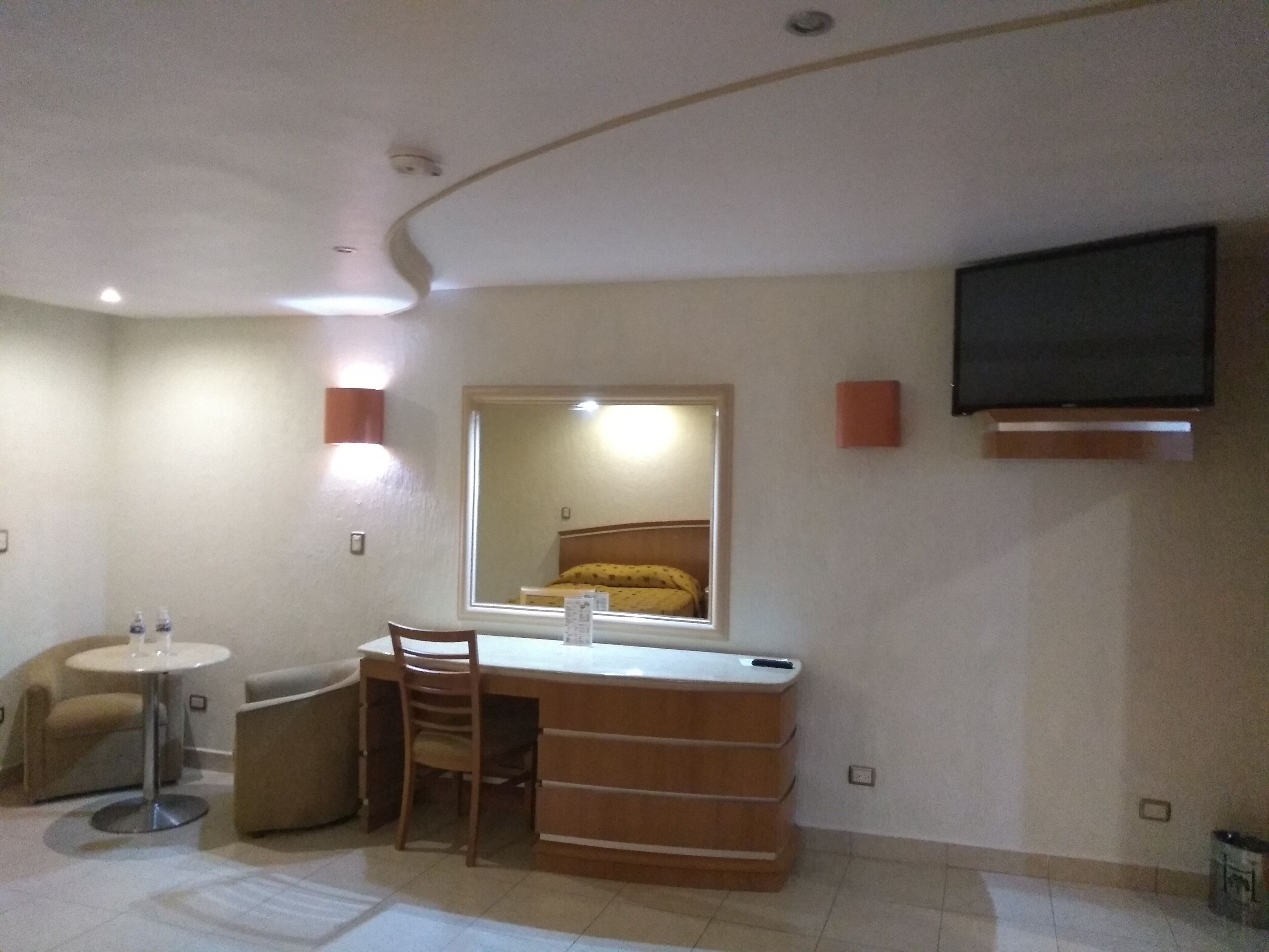 Habitación de hotel con cama, escritorio, silla, mesita redonda, espejo, televisor de pantalla plana montado en la pared y lámpara de pared.