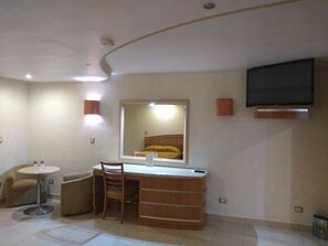 Habitación de hotel con cama, escritorio, silla, mesita redonda, espejo, televisor de pantalla plana montado en la pared y lámpara de pared.