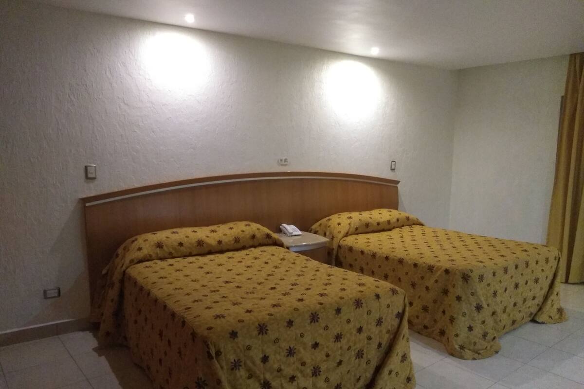 A hotel room with two beds, a wooden headboard, a bedside table with a phone, and a curtain on the window.