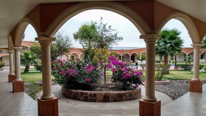A covered courtyard with a central garden, stone planters, and a view of a building with arches in the background. - Hotel Jimenez Plaza (Jimenez)