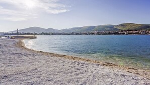 Ein Strand mit klarem Wasser, Kiesufer und einer in der Ferne liegenden Stadt mit Bergen.