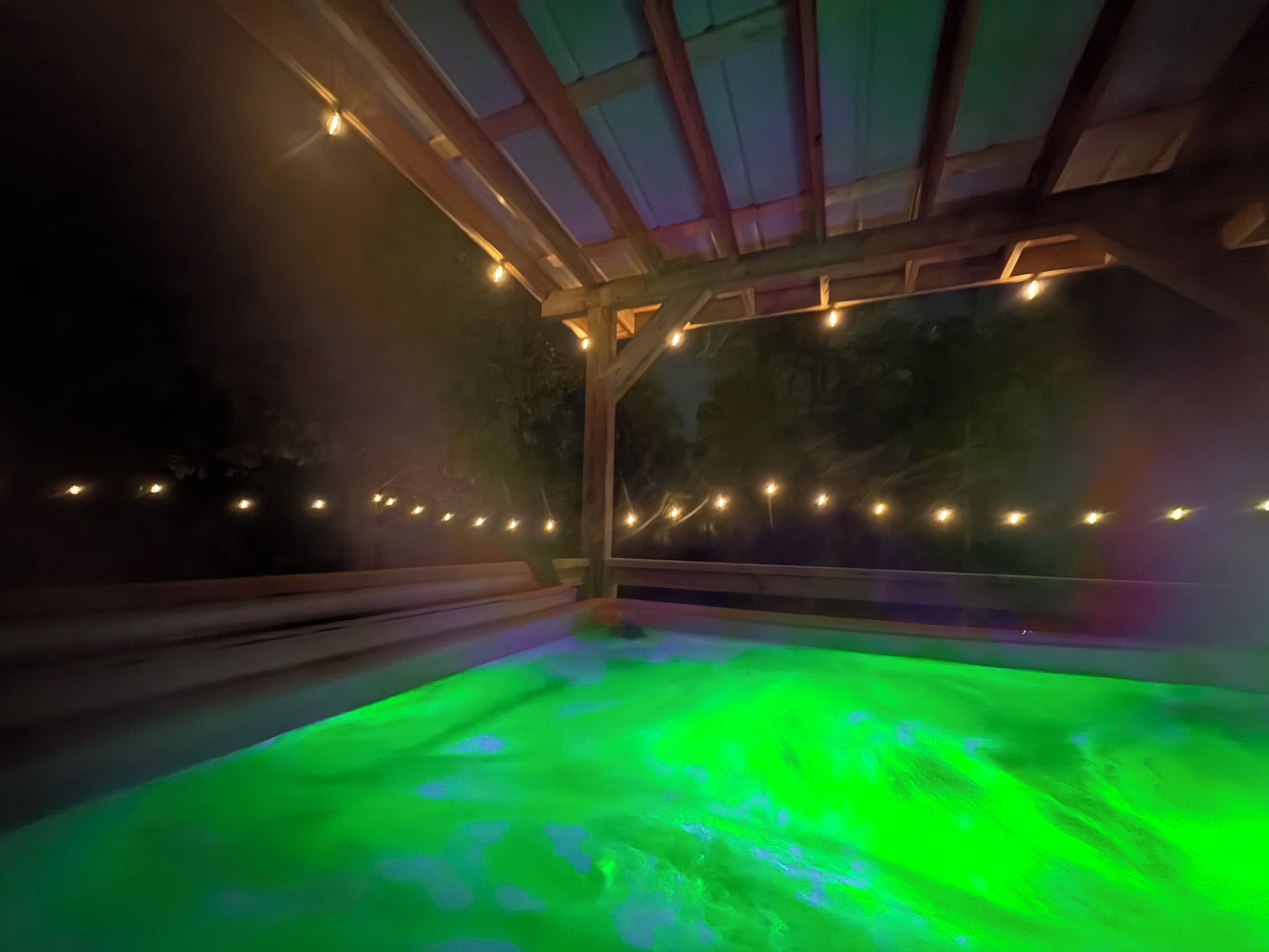 Un jacuzzi con iluminación verde, rodeado por una estructura de madera con luces de cuerda.