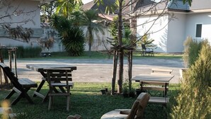 Un espace patio aménagé avec du mobilier en bois, comprenant une table et des chaises, entouré de verdure et offrant une vue sur une route et des bâtiments en arrière-plan.