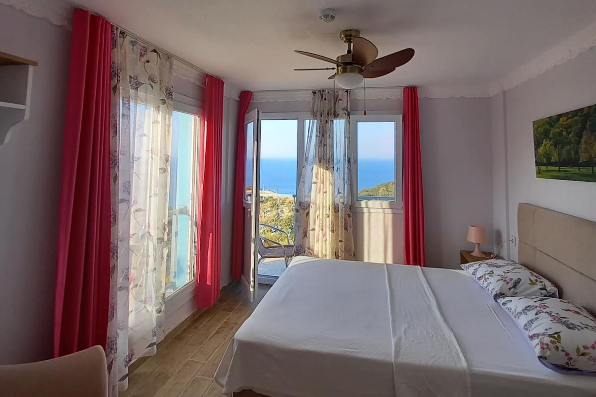 Une chambre avec un lit, une chaise, un ventilateur de plafond et une vue sur la mer.
