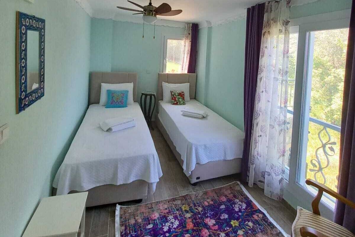 Une chambre avec deux lits simples, un ventilateur de plafond, une petite table, une chaise et un tapis coloré.