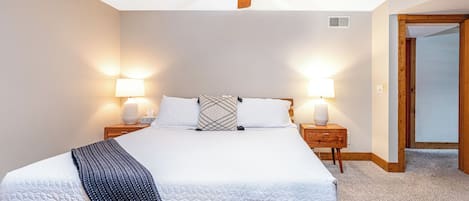 A bedroom with a large bed, two bedside tables with lamps, a ceiling fan, and a door leading to another room.