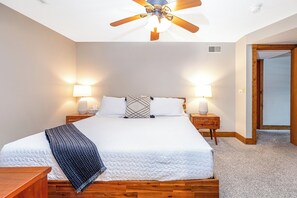 Une chambre à coucher avec un grand lit, deux tables de chevet avec des lampes, un ventilateur de plafond et une porte donnant sur une autre pièce.