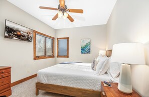 A bedroom with a bed, a nightstand, a lamp, a ceiling fan, and two windows with blinds.