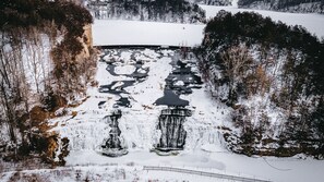A snow-covered landscape with a frozen waterfall, surrounded by trees and a distant building.