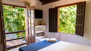 Un dormitorio con cama, televisor montado en la pared, ventana con contraventanas y vista a la vegetación.