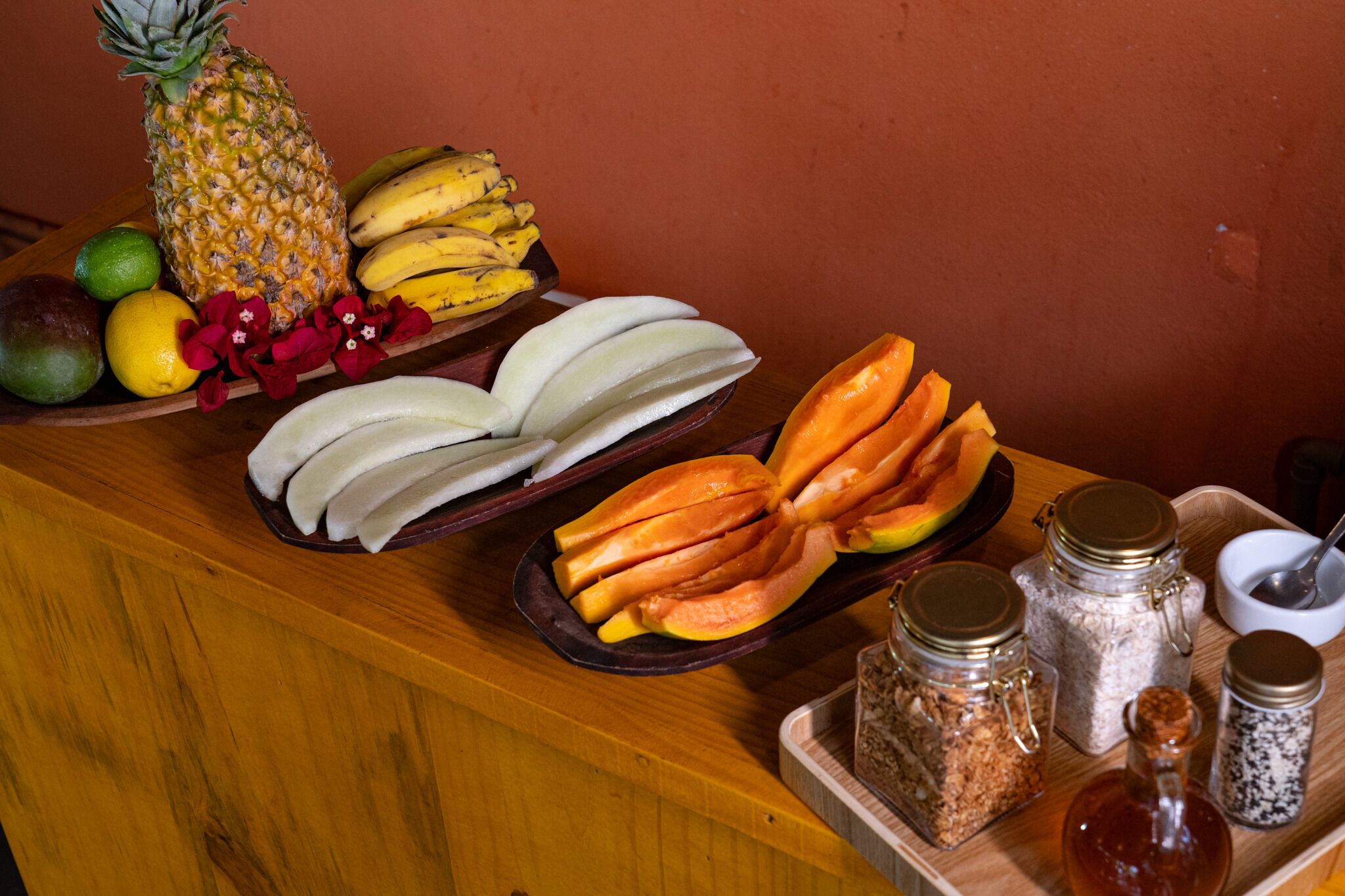 Mesa de buffet de madeira com diversas frutas, incluindo abacaxi, bananas, manga e mamão, juntamente com flores e especiarias.