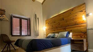 A bedroom with a wooden headboard, a bed with blue bedding, a chair, a window with blinds, and a bedside table. - Rural house Las Gavias  (Matapozuelos)