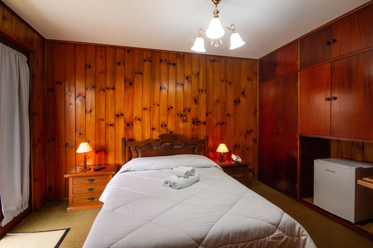 A wooden-paneled room with a bed, two bedside tables with lamps, a nightstand, and a small refrigerator.