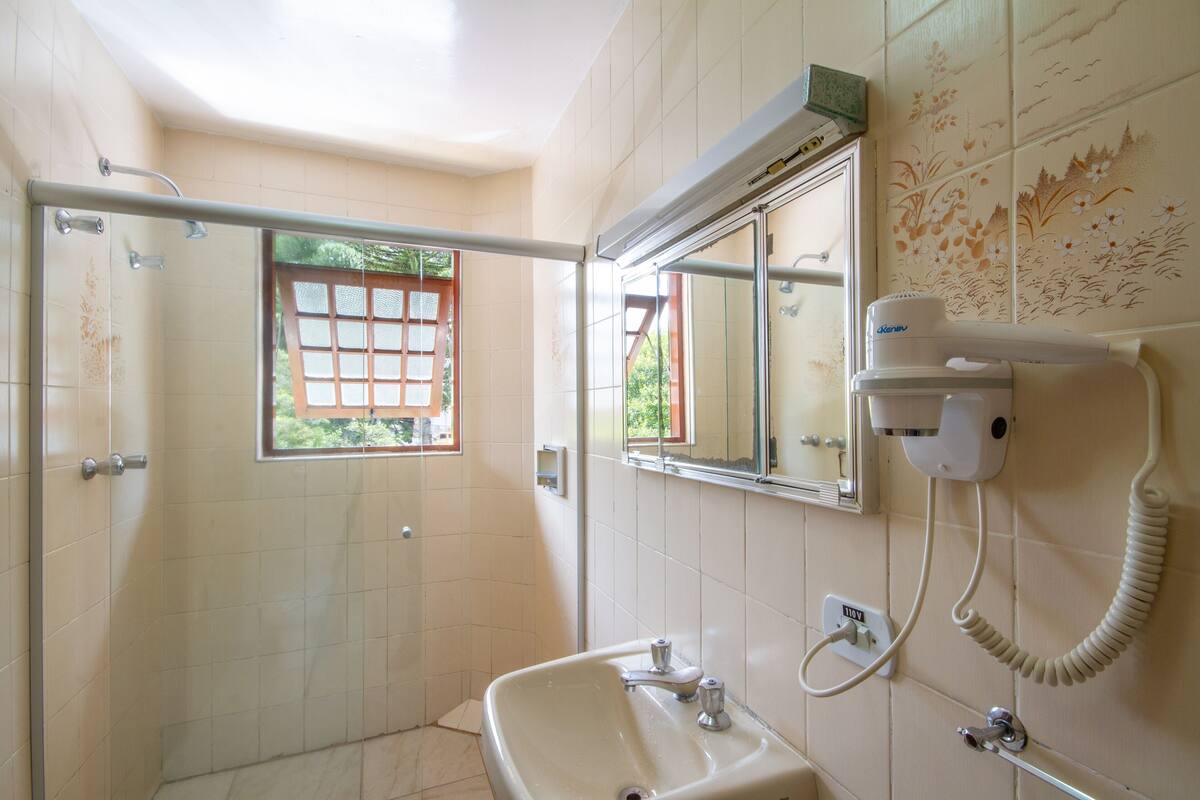 A bathroom with a shower, a sink, a hairdryer, and a window with a view of greenery.