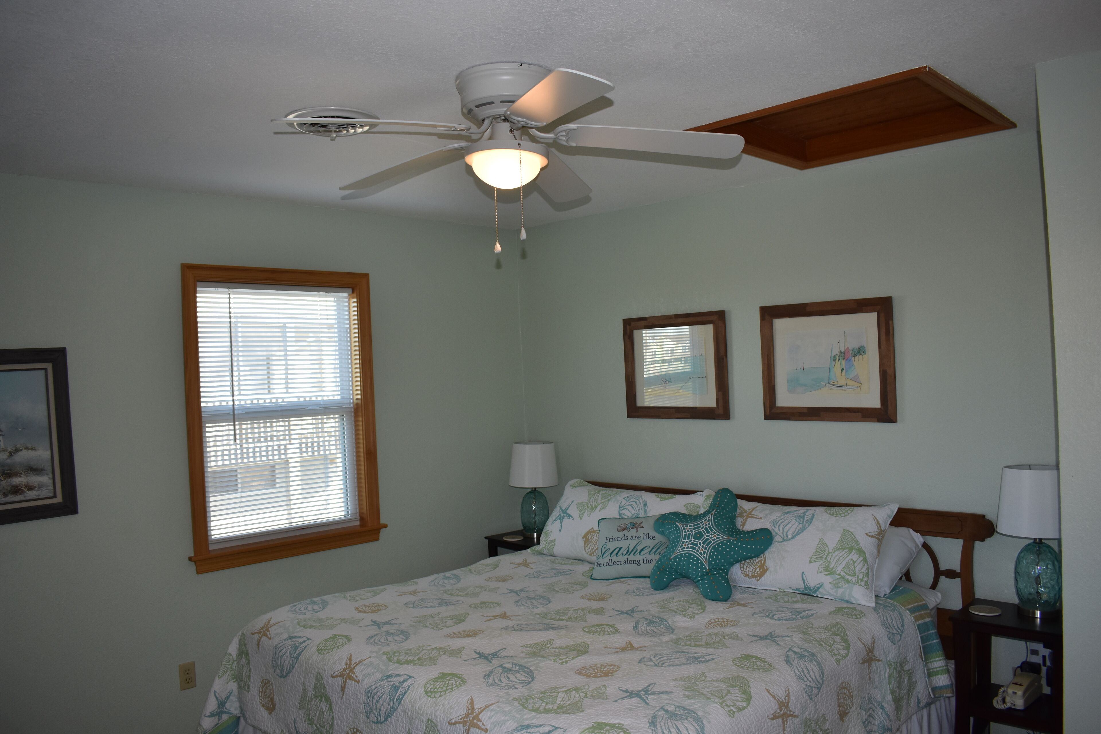 A bedroom with a bed, a ceiling fan, a window with blinds, two framed pictures on the wall, and a nightstand with a lamp.