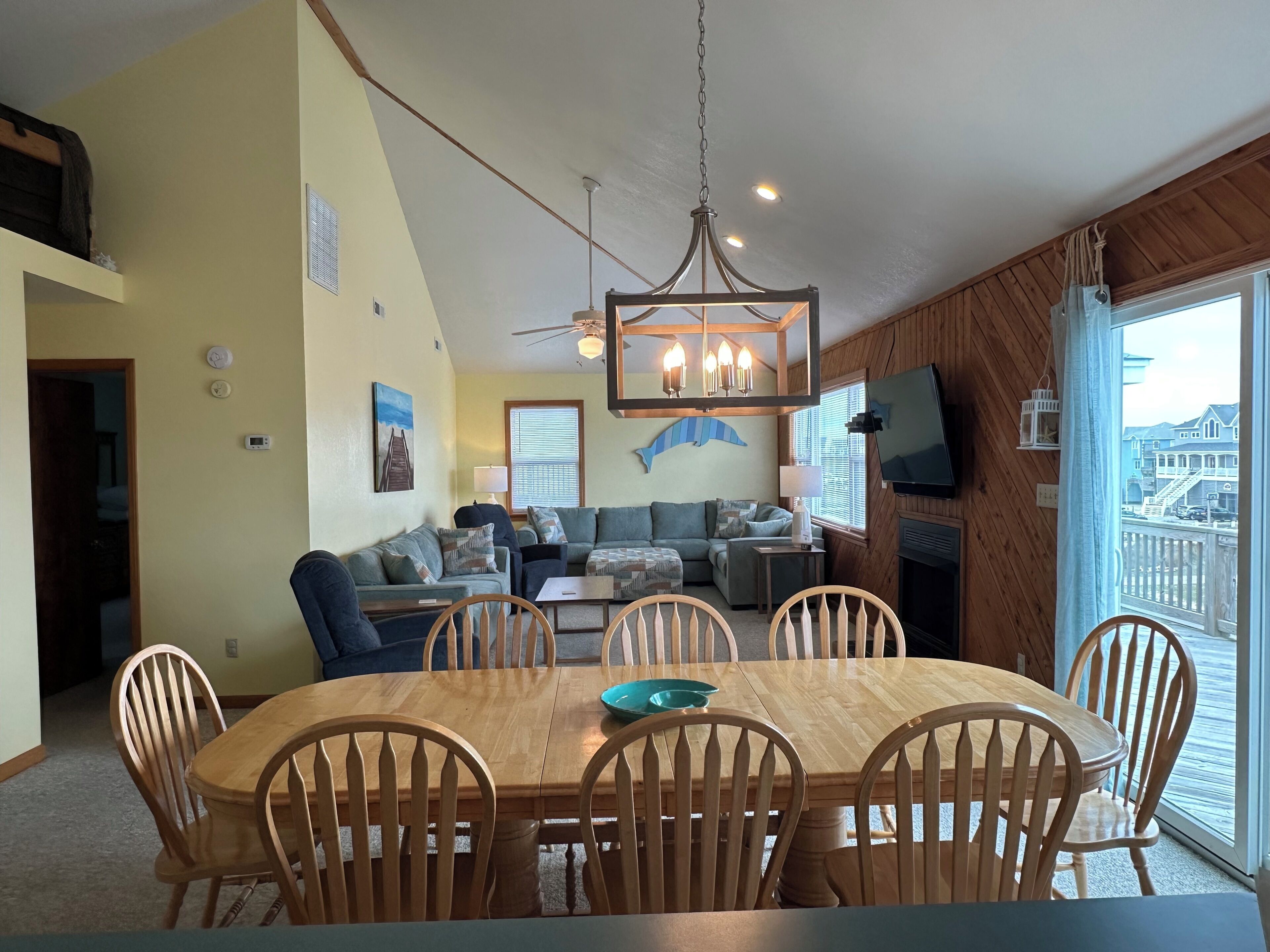 A dining area with a wooden table and chairs, a ceiling fan, and a large window with a view of houses.