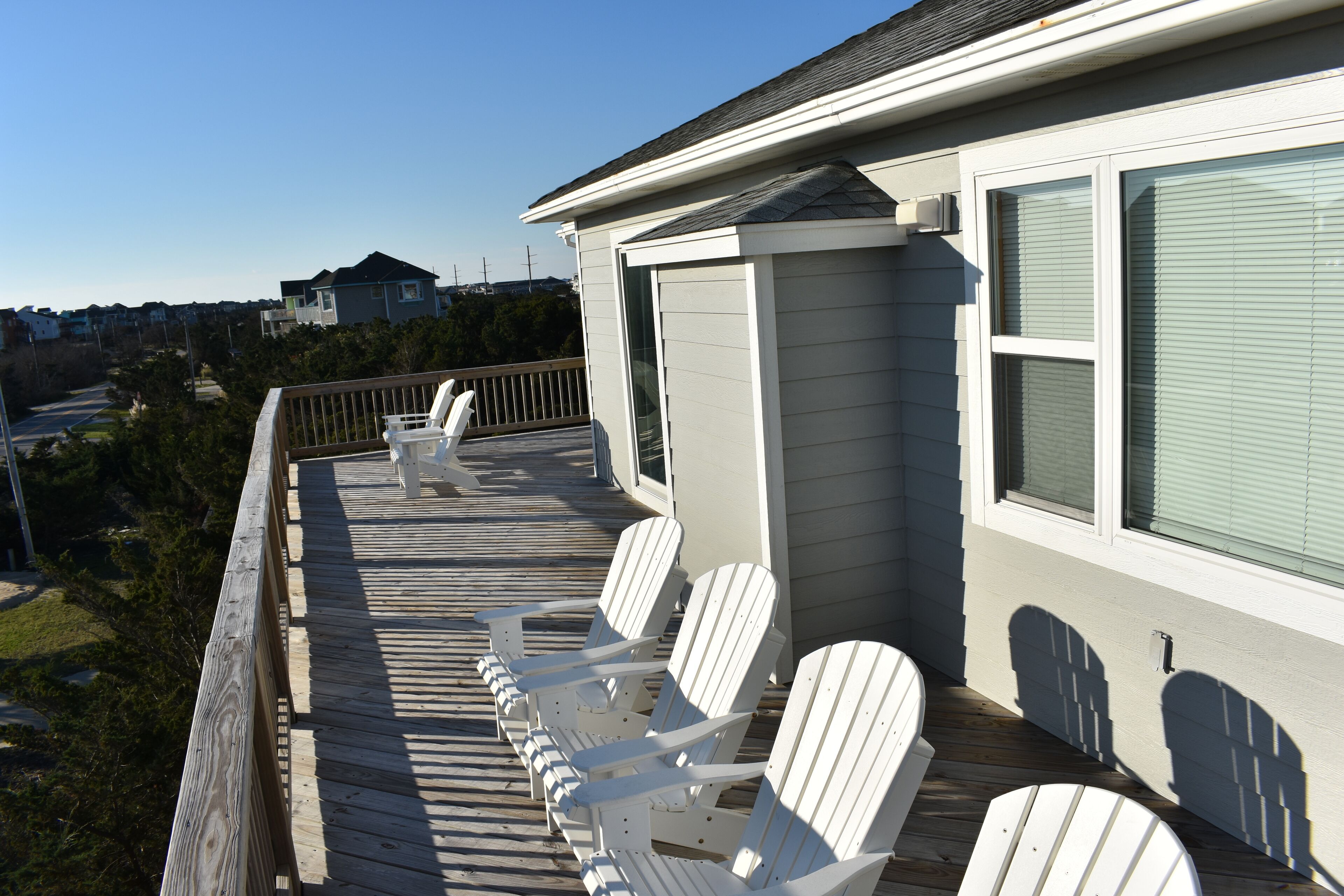 Terraza de madera con sillas Adirondack blancas y una mesa, con vista a una zona residencial.