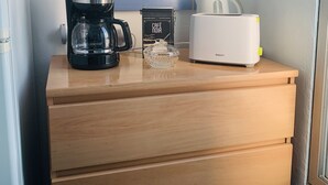 Un aparador de madera con una cafetera, un pequeño recipiente de vidrio y un electrodoméstico blanco, posiblemente un calentador de alimentos, frente a una pared con un espejo que refleja una escalera.