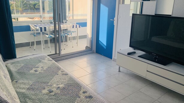 Una sala moderna con un televisor de pantalla plana grande, un ventanal corredero que da a un balcón exterior con una mesa y sillas, y una alfombra estampada en el suelo.