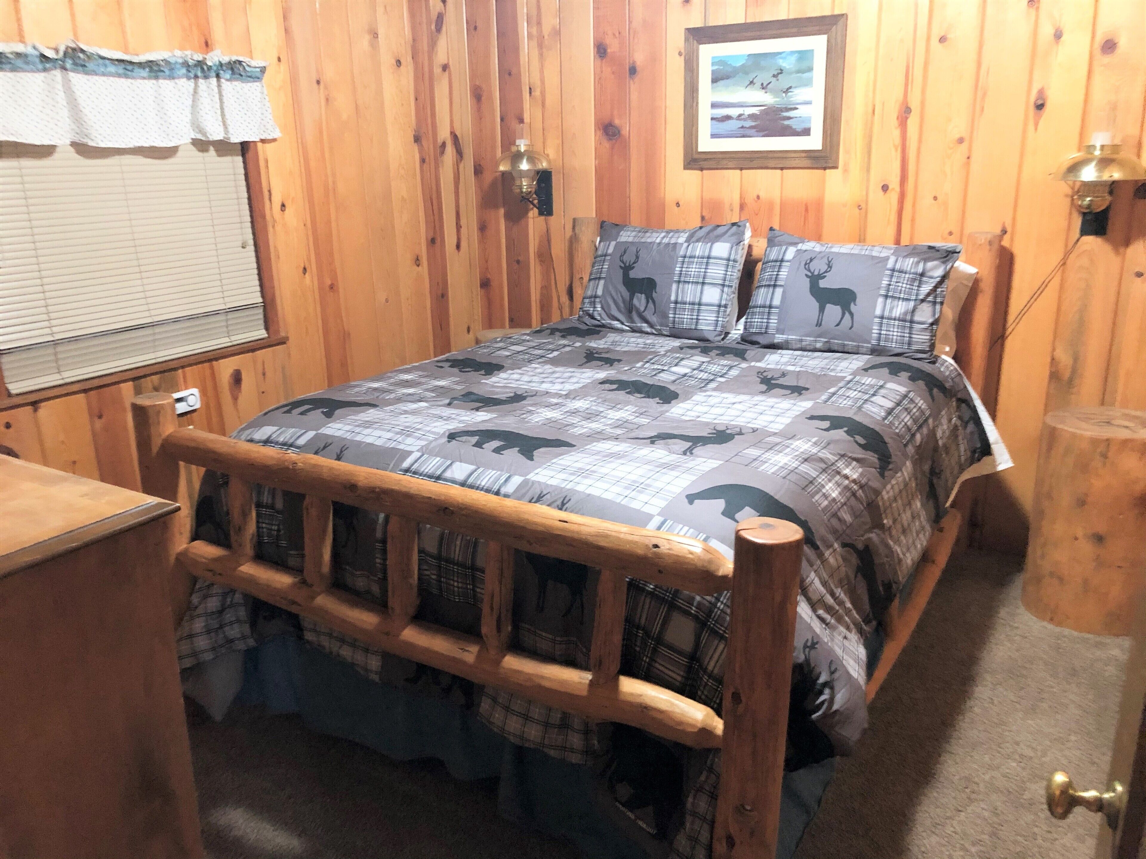 A wooden-paneled room with a bed featuring a plaid bedding set with animal prints, a wooden headboard, a window with blinds, and a framed picture on the wall.