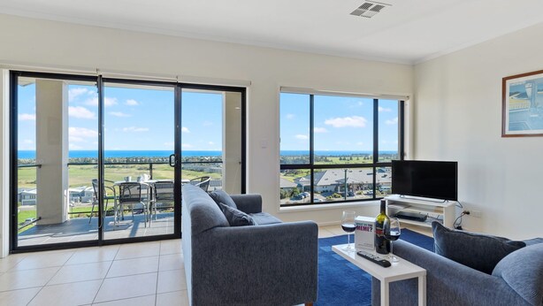 A spacious living room with a large sliding glass door leading to a balcony, a flat-screen TV, and a small table with wine glasses and a bottle.