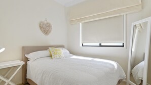 Een slaapkamer met een bed, een nachtkastje, een macramé decoratie aan de muur, een raam met een zonwering en een spiegel.