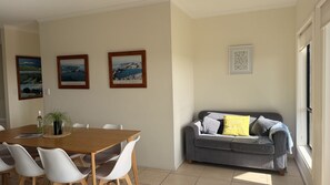 Ein Essbereich mit einem Holztisch und weißen Stühlen, ein graues Sofa mit Kissen sowie gerahmte Landschaftsbilder an der Wand.