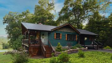 A rustic cabin with a wooden deck, stone foundation, and a metal roof, surrounded by lush greenery and trees.