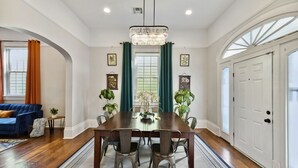 A dining room with a wooden table, chairs, and a chandelier. There are potted plants and artwork on the walls. - The Big Blue House (New Orleans)