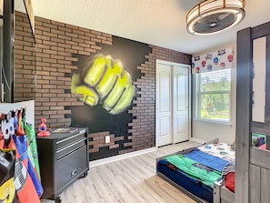 A bedroom with a brick-patterned wall, a bed with a colorful bedspread, a dresser, and a wall-mounted fan. - Disney 8BR Villa • 2 Theaters • Arcade • Pool/Spa (Kissimmee)