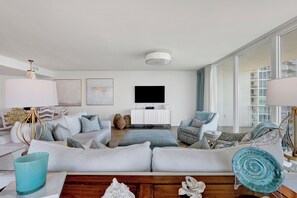 A modern living room with a sectional sofa, a flat-screen TV, and a large window offering a view of a building with balconies. - Modern Style Meets Coastal Chic in this Beautiful Spacious Unit! Complimentary Boat Slip! (Orange Beach)