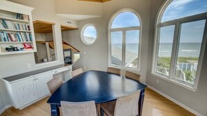 A spacious dining area with a large table and chairs, a kitchen with white cabinets, and large windows offering ocean views. - Duck Sea Sun | Oceanfront | Duck (Duck)