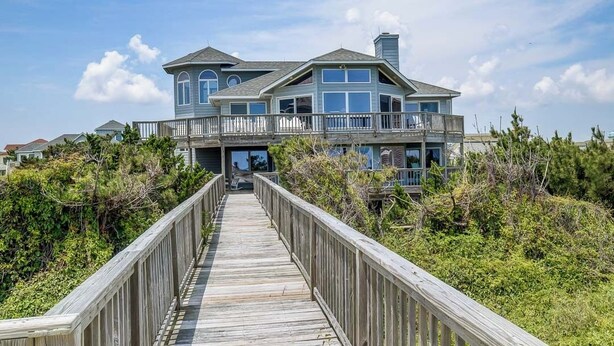 A large, two-story house with a balcony, a wooden deck leading to the entrance, and a backdrop of greenery. - Duck Sea Sun | Oceanfront | Duck (Duck)
