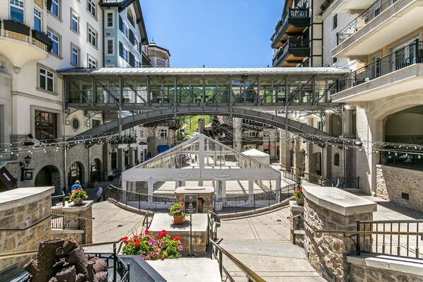 A covered pedestrian bridge with a glass canopy, stone walls, and a central open area with potted plants.