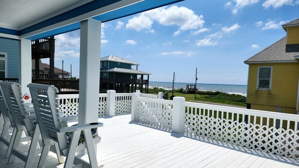 Una terraza de madera con barandas blancas que da a una playa, con casas al fondo.