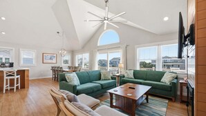 A spacious living area with a high ceiling, a dining area, and large windows offering views of the surroundings. - 1517 - Drift Inn - Steps to Beach | Pool, Hot Tub & Pet Friendly (Corolla)