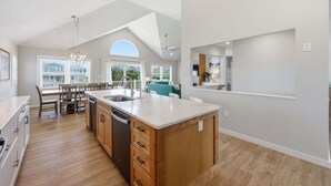 A modern kitchen with a central island, wooden cabinets, and a dining area with chairs. - 1517 - Drift Inn - Steps to Beach | Pool, Hot Tub & Pet Friendly (Corolla)