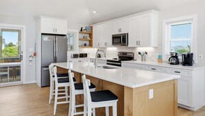 A modern kitchen with white cabinetry, a central island with bar stools, a stainless steel refrigerator, a microwave, and a coffee maker. - 1517 - Drift Inn - Steps to Beach | Pool, Hot Tub & Pet Friendly (Corolla)