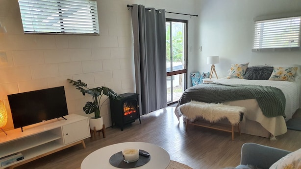 A modern living room with a flat-screen TV, a small table with a bowl, a sofa, a bed with a green blanket, a small heater, and a potted plant. - Farm Stay book  a Private Cabin - Relax Breathe Walk (Palmwoods)