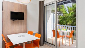Een moderne kamer met een witte tafel en oranje stoelen, een flatscreen televisie bevestigd aan een houten paneel, en een balkon met een witte reling en tuinmeubilair.